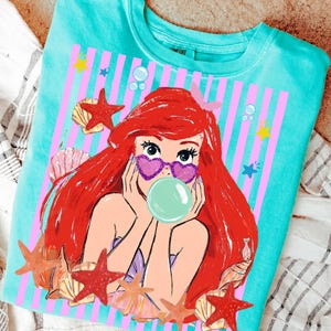 May include: Turquoise crew neck sweatshirt with a mermaid graphic. The mermaid has red hair, heart-shaped sunglasses, and a bubble. The design includes pink stripes, starfish, seashells, and bubbles, creating a whimsical, beachy theme.