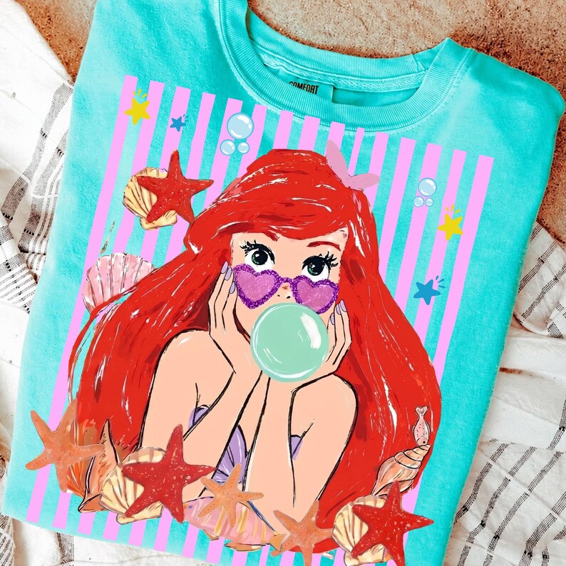 May include: Turquoise crew neck sweatshirt with a mermaid graphic. The mermaid has red hair, heart-shaped sunglasses, and a bubble. The design includes pink stripes, starfish, seashells, and bubbles, creating a whimsical, beachy theme.