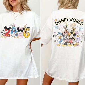 Może przedstawiać: Biała koszulka z postaciami Disneya i napisem "2026" z przodu. Tył koszulki przedstawia "Disney World" z r&oacute;żnymi postaciami i rokiem "1971". Projekt zawiera Myszkę Miki, Minnie, Kaczora Donalda i Goofy'ego.