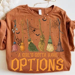 May include: Burnt orange crew neck sweatshirt featuring a graphic of several witch brooms in various colors and styles. The text "A Girl's Gotta Have Options" is printed in orange block letters below the brooms. Bats and spiderwebs are also included.