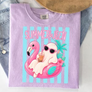 K&ouml;nnte beinhalten: Helllila T-Shirt mit dem Wort "SUMMERWEEN" in Pink und einem Cartoon-Geist mit Sonnenbrille, der auf einem rosa Flamingo-Schwimmer treibt. Der Geist h&auml;lt ein Getr&auml;nk mit Boba-Perlen.