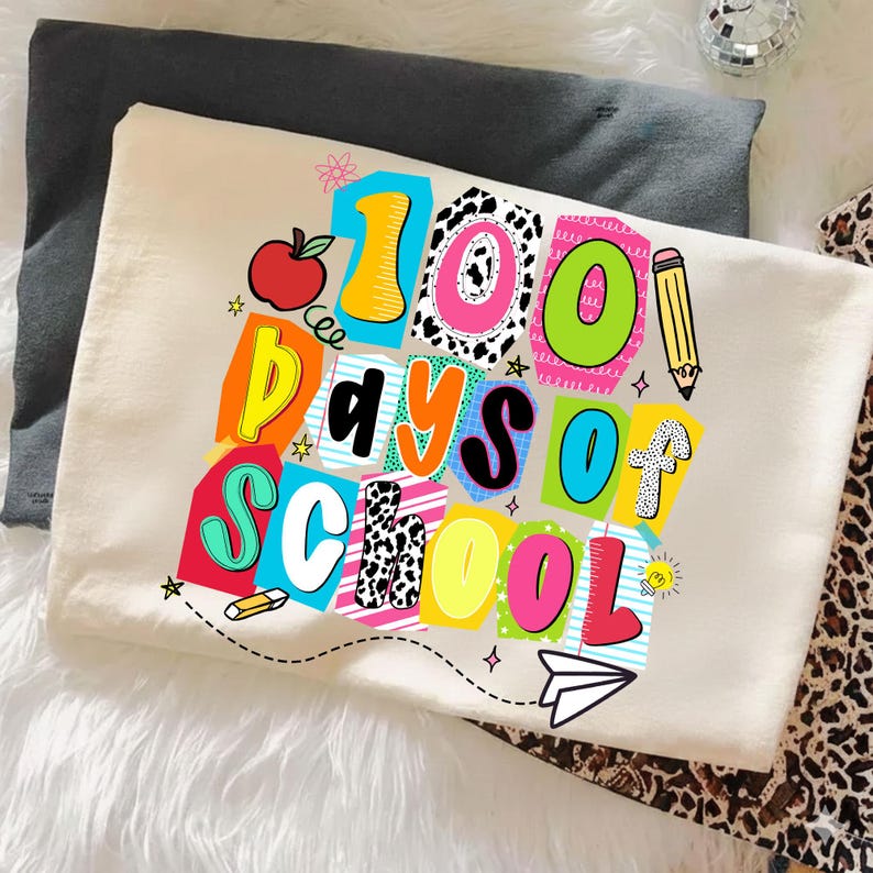 May include: A cream-colored t-shirt with the text "100 Days of School" in colorful, block letters. The design includes an apple, pencil, and paper airplane. The shirt is folded on a white surface.