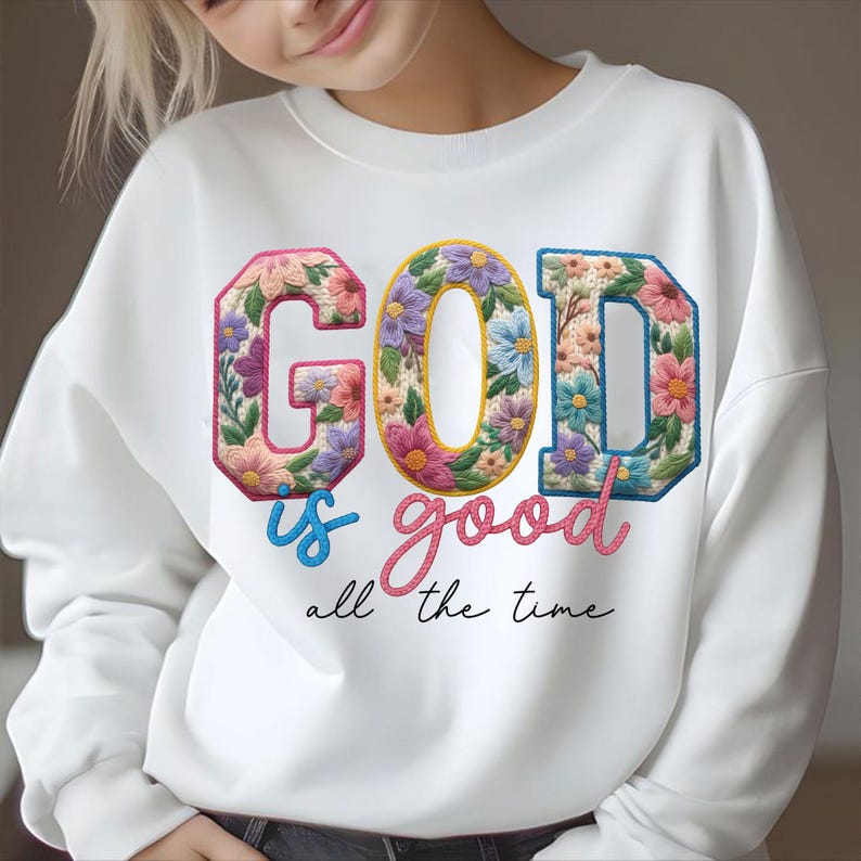 Op de afbeelding: Witte sweatshirt met de tekst "GOD is good all the time". De letters zijn versierd met bloemmotieven in roze, paars, geel en blauw. De woorden "is" en "good" zijn in script geschreven, respectievelijk in blauw en roze.