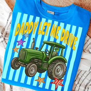 Pu&ograve; includere: Felpa blu con grafica di un trattore verde e la scritta gialla "DADDY LET ME DRIVE". Il design include strisce blu e bianche e stelle colorate.