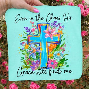 May include: A light blue square fabric with the text "Even in the Chaos His Grace still finds me." A large, colorful cross is the central design, surrounded by vibrant flowers in pink, orange, purple, and blue.