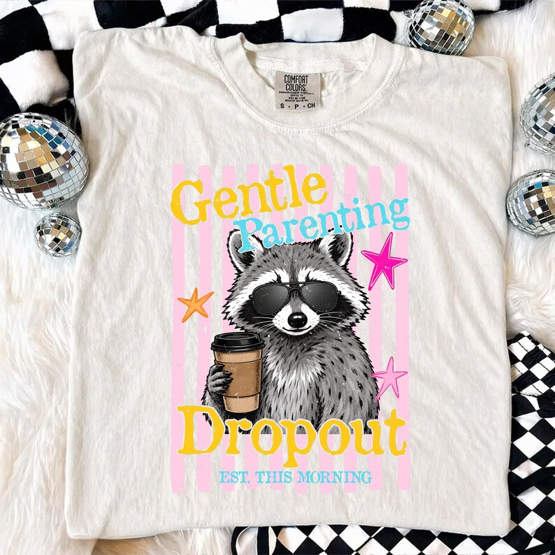 Op de afbeelding: Cr&egrave;mekleurig T-shirt met een afbeelding van een wasbeer met zonnebril en een koffiekopje. De tekst luidt "Gentle Parenting Dropout" met steraccenten. Het shirt is van het merk Comfort Colors.
