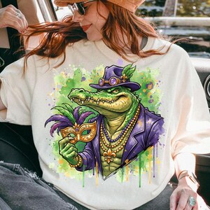 May include: Cream-colored sweatshirt featuring a vibrant illustration of an alligator in a purple suit and hat, holding a gold and purple mask. The design includes a beaded necklace and a fleur-de-lis, with green and purple watercolor accents.