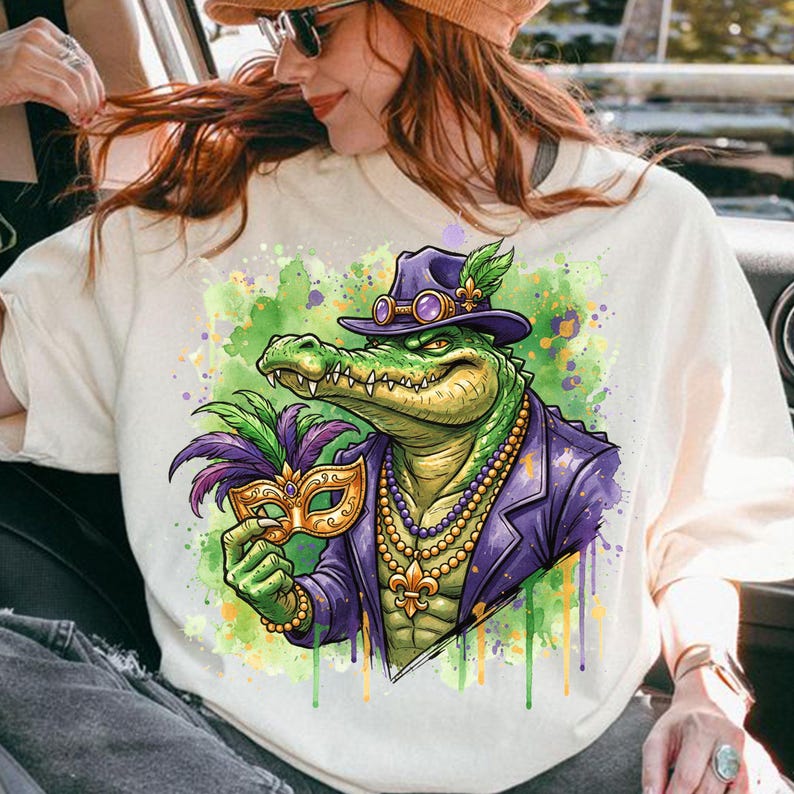 May include: Cream-colored sweatshirt featuring a vibrant illustration of an alligator in a purple suit and hat, holding a gold and purple mask. The design includes a beaded necklace and a fleur-de-lis, with green and purple watercolor accents.