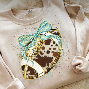 May include: A beige sweatshirt featuring a football design with a cowhide pattern and a light blue bow. The football has white laces and gold accents. The design is embellished with gold and blue speckles.