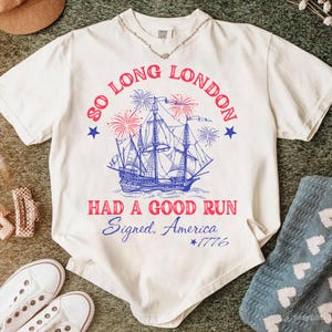 May include: Cream-colored t-shirt with a blue and red graphic of a ship, fireworks, and text that reads "SO LONG LONDON HAD A GOOD RUN Signed, America *1776". The shirt has a V-neck and short sleeves.