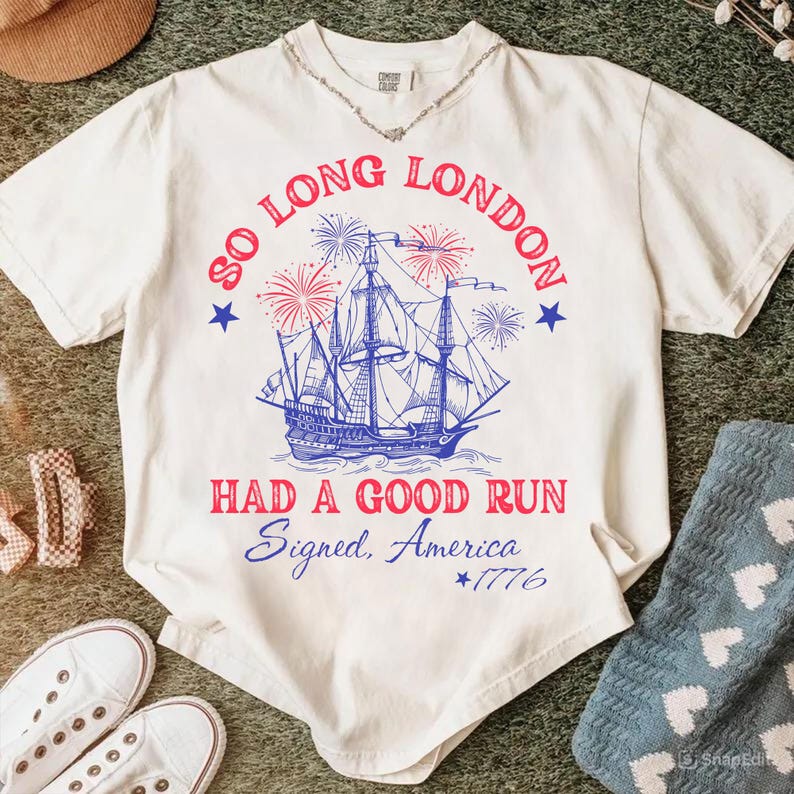 May include: Cream-colored t-shirt with a blue and red graphic of a ship, fireworks, and text that reads "SO LONG LONDON HAD A GOOD RUN Signed, America *1776". The shirt has a V-neck and short sleeves.