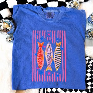 Pode incluir: Camiseta azul Comfort Colors com um gr&aacute;fico colorido de tr&ecirc;s peixes. Os peixes s&atilde;o em tons de vermelho, rosa e amarelo, com listras verticais rosa e estrelas azuis. A camisa tem gola redonda e o logotipo Comfort Colors.