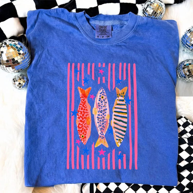 Pode incluir: Camiseta azul Comfort Colors com um gr&aacute;fico colorido de tr&ecirc;s peixes. Os peixes s&atilde;o em tons de vermelho, rosa e amarelo, com listras verticais rosa e estrelas azuis. A camisa tem gola redonda e o logotipo Comfort Colors.