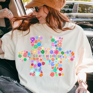 May include: Cream-colored t-shirt featuring a colorful floral puzzle piece design. The design includes various flowers in shades of pink, blue, yellow, and green. The text "IT'S OKAY TO BE DIFFERENT" is printed in blue.