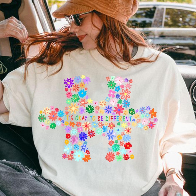 May include: Cream-colored t-shirt featuring a colorful floral puzzle piece design. The design includes various flowers in shades of pink, blue, yellow, and green. The text "IT'S OKAY TO BE DIFFERENT" is printed in blue.