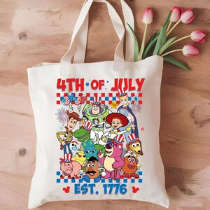 May include: A white canvas tote bag with a 4th of July theme featuring characters from the Toy Story franchise. The bag has the text "4th of July" and "Est. 1776" printed on it. The characters are wearing red, white, and blue patriotic hats and clothing.