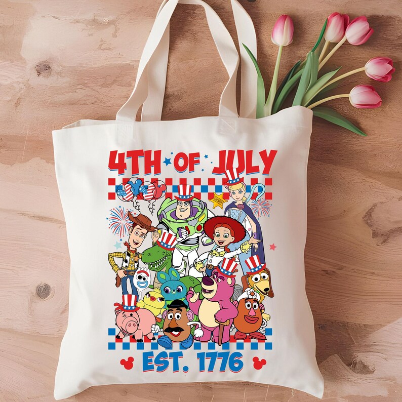 May include: A white canvas tote bag with a 4th of July theme featuring characters from the Toy Story franchise. The bag has the text "4th of July" and "Est. 1776" printed on it. The characters are wearing red, white, and blue patriotic hats and clothing.