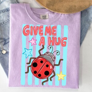 Pu&ograve; includere: T-shirt lilla chiaro con la scritta "GIVE ME A HUG" in rosso, con un'illustrazione di coccinella. Il design include strisce verticali blu, una stella rosa, una stella blu e una stella gialla.