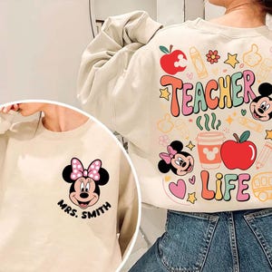May include: Cream-coloured sweatshirt with a Minnie Mouse graphic and the text "Mrs. Smith" on the front. The back features colourful graphics with the words "Teacher Life" and images of apples, pencils, and Mickey Mouse.