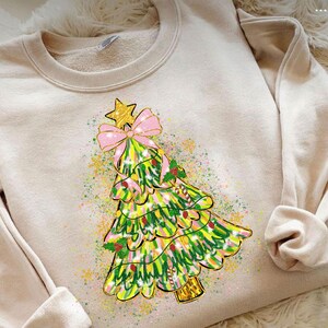 May include: A beige sweatshirt featuring a colorful Christmas tree design. The tree is decorated with a pink bow, gold star, and holly berries. The design has a watercolor effect with green, yellow, and pink hues.