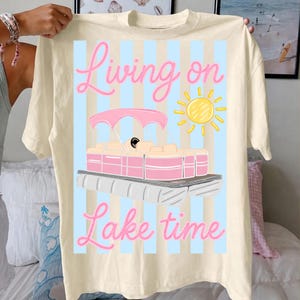 May include: Cream-colored t-shirt with a graphic of a pink and white pontoon boat, a yellow sun, and the words "Living on Lake time" in pink script. The design is set against a light blue striped background.