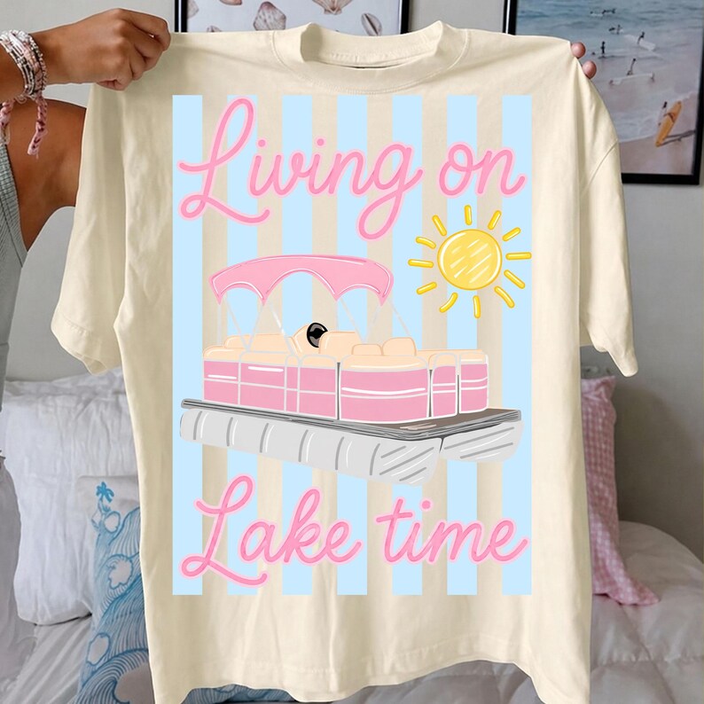 May include: Cream-colored t-shirt with a graphic of a pink and white pontoon boat, a yellow sun, and the words "Living on Lake time" in pink script. The design is set against a light blue striped background.