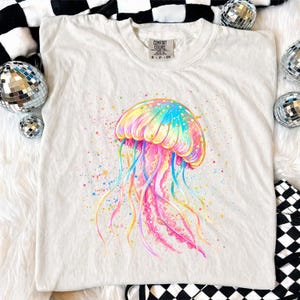 May include: A white Comfort Colors t-shirt featuring a vibrant watercolor jellyfish design. The jellyfish is depicted in shades of pink, yellow, blue, and orange, with colorful paint splatters around it. The shirt has a crew neck and short sleeves.