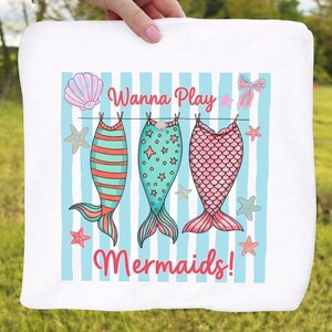 May include: White towel with a design featuring three mermaid tails hanging on a clothesline. The tails are in shades of teal, pink, and coral, with the words "Wanna Play Mermaids!" in pink.