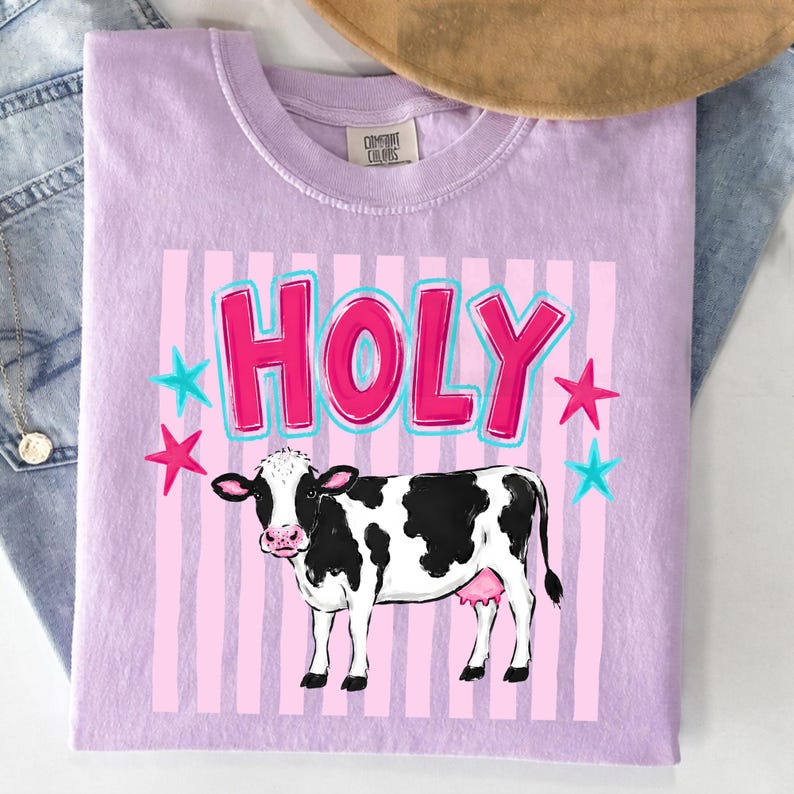 Puede incluir: Una camiseta de color lila claro con la palabra "HOLY" en letras rosas con contorno azul. Debajo del texto hay una ilustraci&oacute;n de una vaca en blanco y negro. Estrellas rosas y azules salpican el dise&ntilde;o.