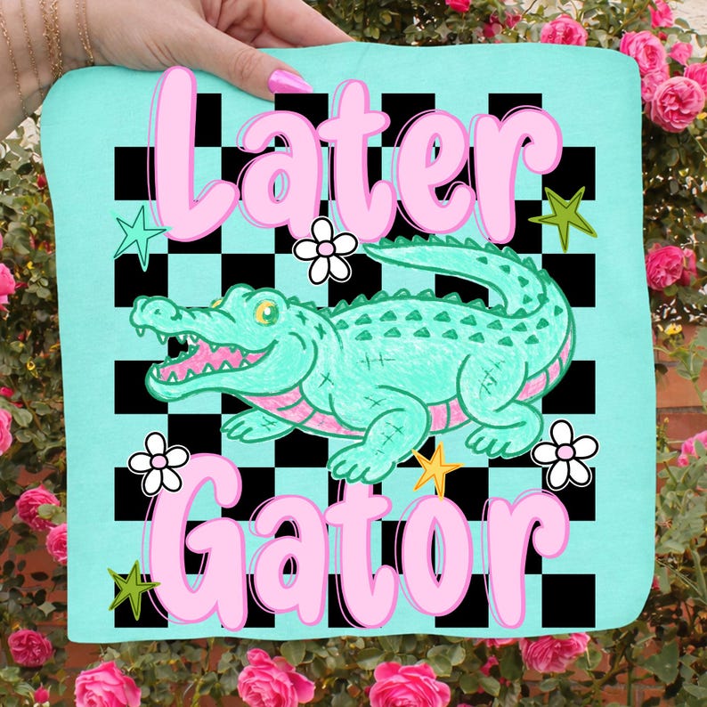 May include: A light blue square textile with the words "Later Gator" in pink, featuring a cartoon alligator. The design includes a black and white checkered background, flowers, and stars.