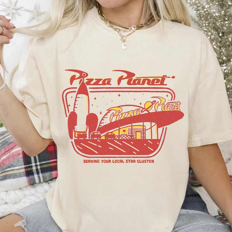 Puede incluir: Camiseta color crema con un dise&ntilde;o gr&aacute;fico rojo y amarillo del logo de Pizza Planet, que incluye un cohete y el texto "Pizza Planet" y "Serving Your Local Star Cluster".