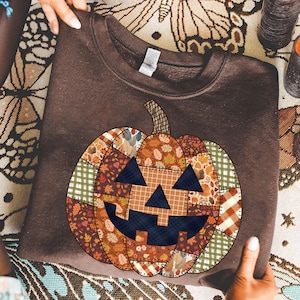 May include: A brown sweatshirt featuring a patchwork pumpkin design. The pumpkin has a smiling face with a dark blue mouth and eyes. The patchwork includes fall leaves, plaid, and other autumn-themed patterns. The sweatshirt is held by two hands.