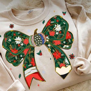 May include: Beige sweatshirt featuring a large, decorative bow design. The bow is green with red poinsettias, white flowers, and gold trim. A pumpkin-shaped center with various patterns adds detail. Red and white ribbon accents complete the festive look.