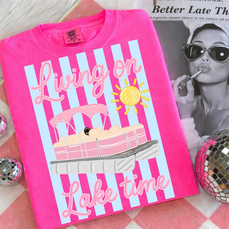 May include: A hot pink sweatshirt with the words "Living on Lake Time" in light pink script. The design includes a pink and cream-colored boat, a yellow sun, and light blue vertical stripes.