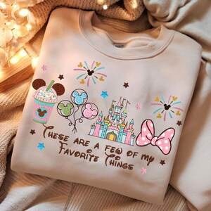 May include: Beige sweatshirt with colorful embroidered Disney-themed design. Features a castle, balloons, bow, fireworks, and a drink with Mickey Mouse ears. Text reads "These are a few of my favorite things."