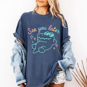 May include: Navy blue t-shirt with the text "See you later" and a cartoon alligator wearing sunglasses. The alligator is outlined in a light blue color with a yellow outline. The text is in pink, yellow, and light blue.