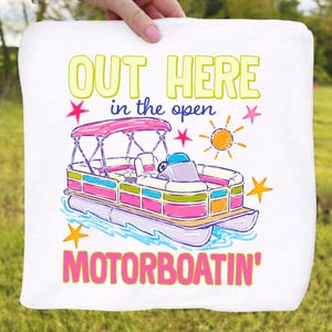 May include: White towel with a colorful illustration of a pontoon boat. The boat has a pink canopy and is surrounded by stars and a sun graphic. Text reads "OUT HERE in the open MOTORBOATING'" in yellow, pink, and blue.