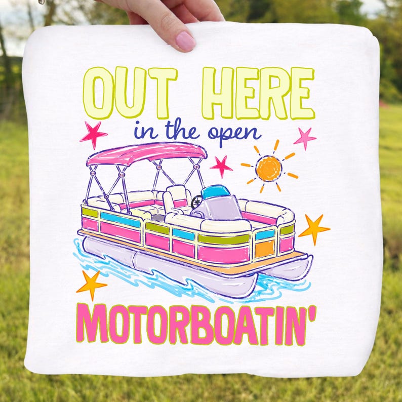 May include: White towel with a colorful illustration of a pontoon boat. The boat has a pink canopy and is surrounded by stars and a sun graphic. Text reads "OUT HERE in the open MOTORBOATING'" in yellow, pink, and blue.