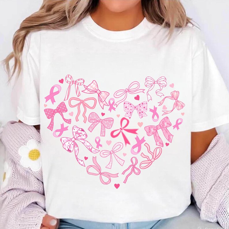 May include: White t-shirt featuring a heart-shaped design composed of pink ribbons and awareness ribbons. The design includes various bow styles and small hearts, all in shades of pink. The shirt is a casual, round-neck style.
