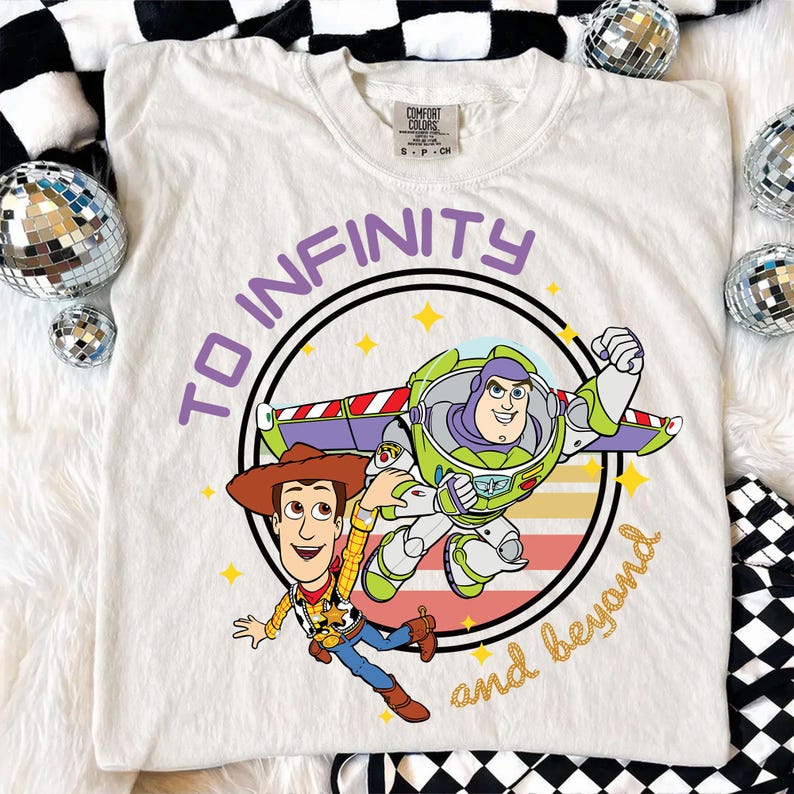 Puede incluir: Camiseta blanca con un dise&ntilde;o de Woody y Buzz Lightyear de Toy Story. El dise&ntilde;o incluye el texto "To Infinity and Beyond" en morado y dorado. La camiseta es de la marca Comfort Colors.