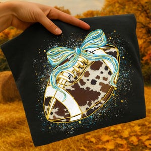 May include: Black t-shirt featuring a football design with a cowhide pattern, white stripes, and gold accents. A blue and gold bow adorns the top of the football. The design is set against a backdrop of autumn colors.