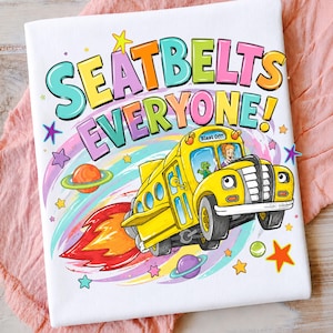 Op de afbeelding: Een wit t-shirt met een kleurrijke afbeelding van een gele schoolbus die door de ruimte vliegt met een raketvlam als uitlaat. De tekst SEATBELTS EVERYONE! staat in grote, veelkleurige letters boven de bus, met een bestuurder en een groen wezen erin.