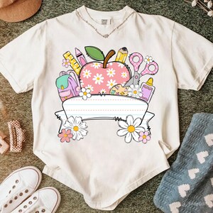 May include: Cream-colored t-shirt featuring a school-themed graphic. The design includes a pink apple with daisy accents, school supplies, and a blank banner. The shirt has a v-neck and short sleeves.