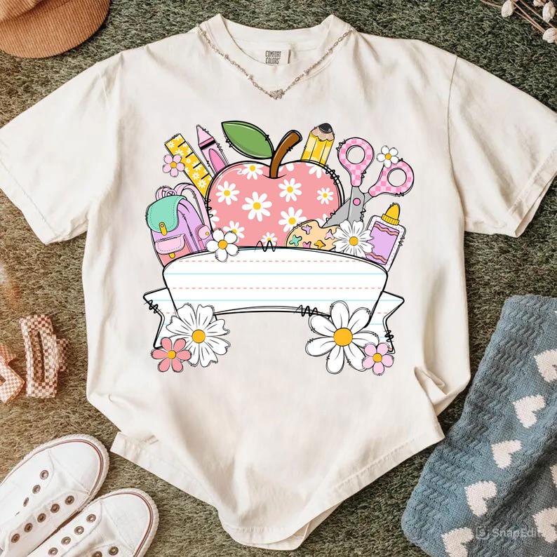 May include: Cream-colored t-shirt featuring a school-themed graphic. The design includes a pink apple with daisy accents, school supplies, and a blank banner. The shirt has a v-neck and short sleeves.