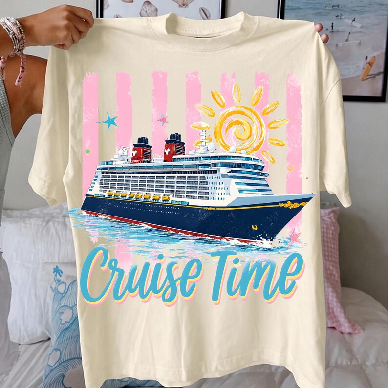 May include: Cream-coloured t-shirt featuring a cruise ship graphic with the text "Cruise Time." The design includes a blue and white ship, a yellow sun, pink stripes, and blue stars. The shirt is held up by a person.