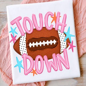 May include: A white t-shirt featuring a brown football graphic with white laces and black polka dot side panels. The text "TOUCH DOWN" is printed in large pink letters above and below the football, surrounded by small orange and blue stars.