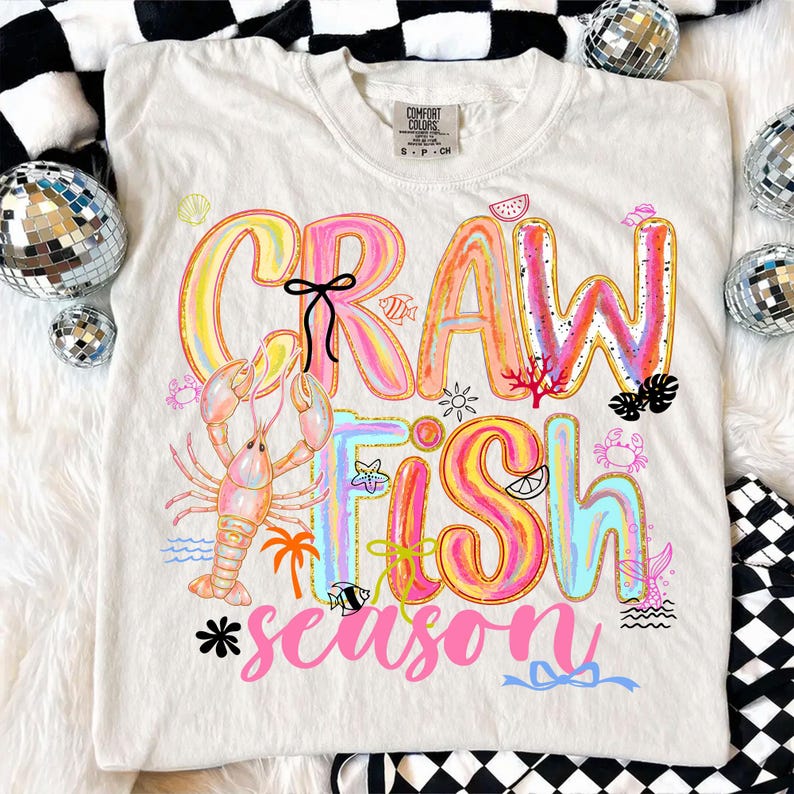 May include: A white Comfort Colors t-shirt with a colorful graphic that says "CRAWFISH SEASON." The design includes a crawfish, palm trees, and other aquatic elements in shades of pink, yellow, and blue.