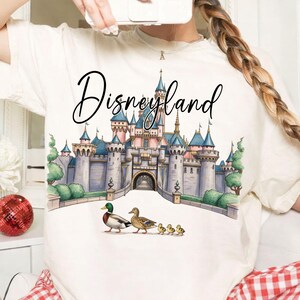 May include: Cream-colored t-shirt featuring a watercolor illustration of Disneyland's castle with blue turrets and pink walls. The word "Disneyland" is written above the castle in a cursive font. A family of ducks walks in front of the castle.