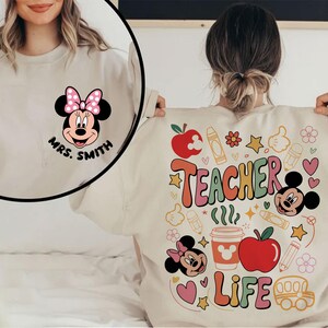 May include: A beige sweatshirt with a Minnie Mouse graphic and the text "Mrs. Smith" on the front. The back displays a colourful design with "Teacher Life" and school-themed illustrations, including apples, crayons, and Mickey Mouse.
