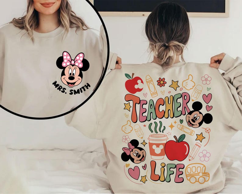 May include: A beige sweatshirt with a Minnie Mouse graphic and the text "Mrs. Smith" on the front. The back displays a colourful design with "Teacher Life" and school-themed illustrations, including apples, crayons, and Mickey Mouse.